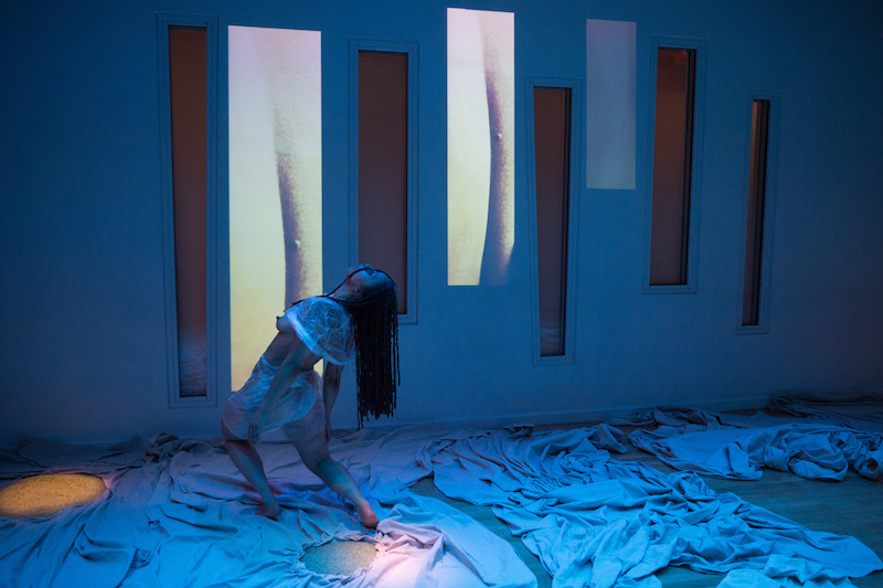 Samantha Speis hinges at her hips. White fabric is strewn across the floor. Two video projections are cast on the wall.